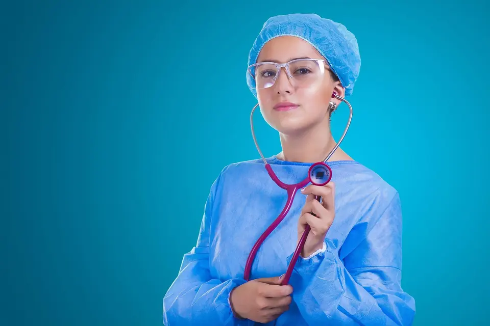 A doctor in blue scrubs and safety glasses holding a stethoscope